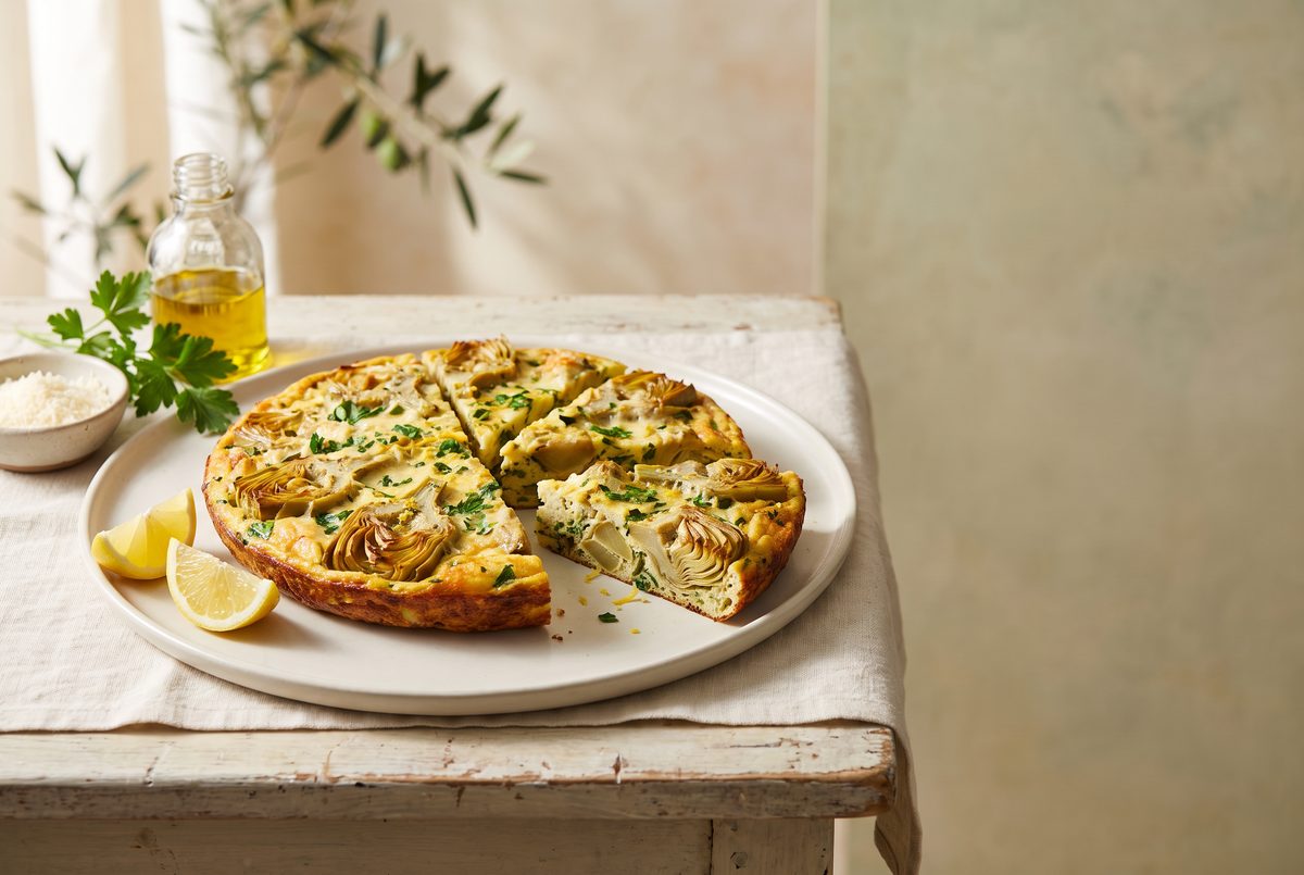 A sliced artichoke frittata on a white plate with lemon wedges, parsley, grated cheese, and olive oil on a rustic wooden table.