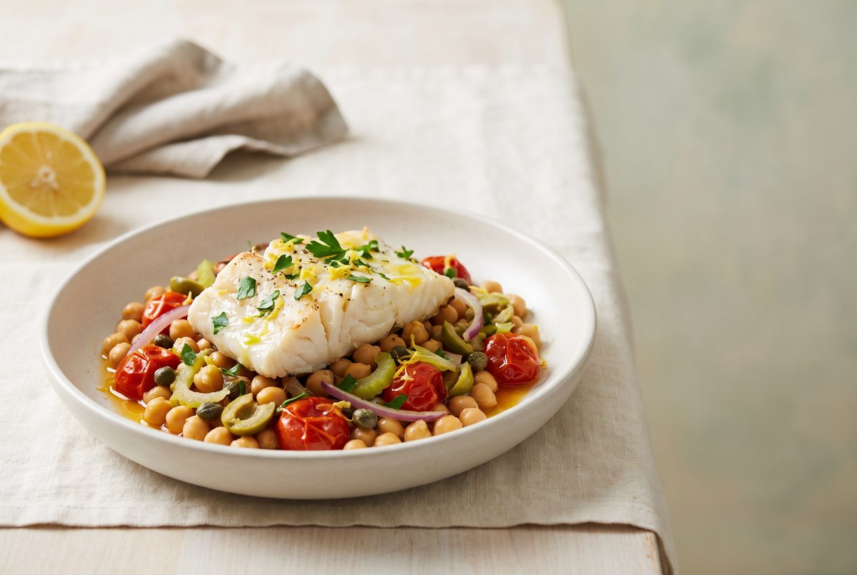 A white fish fillet on chickpeas, roasted tomatoes, olives, red onion, and capers, garnished with parsley and lemon zest, with a lemon.
