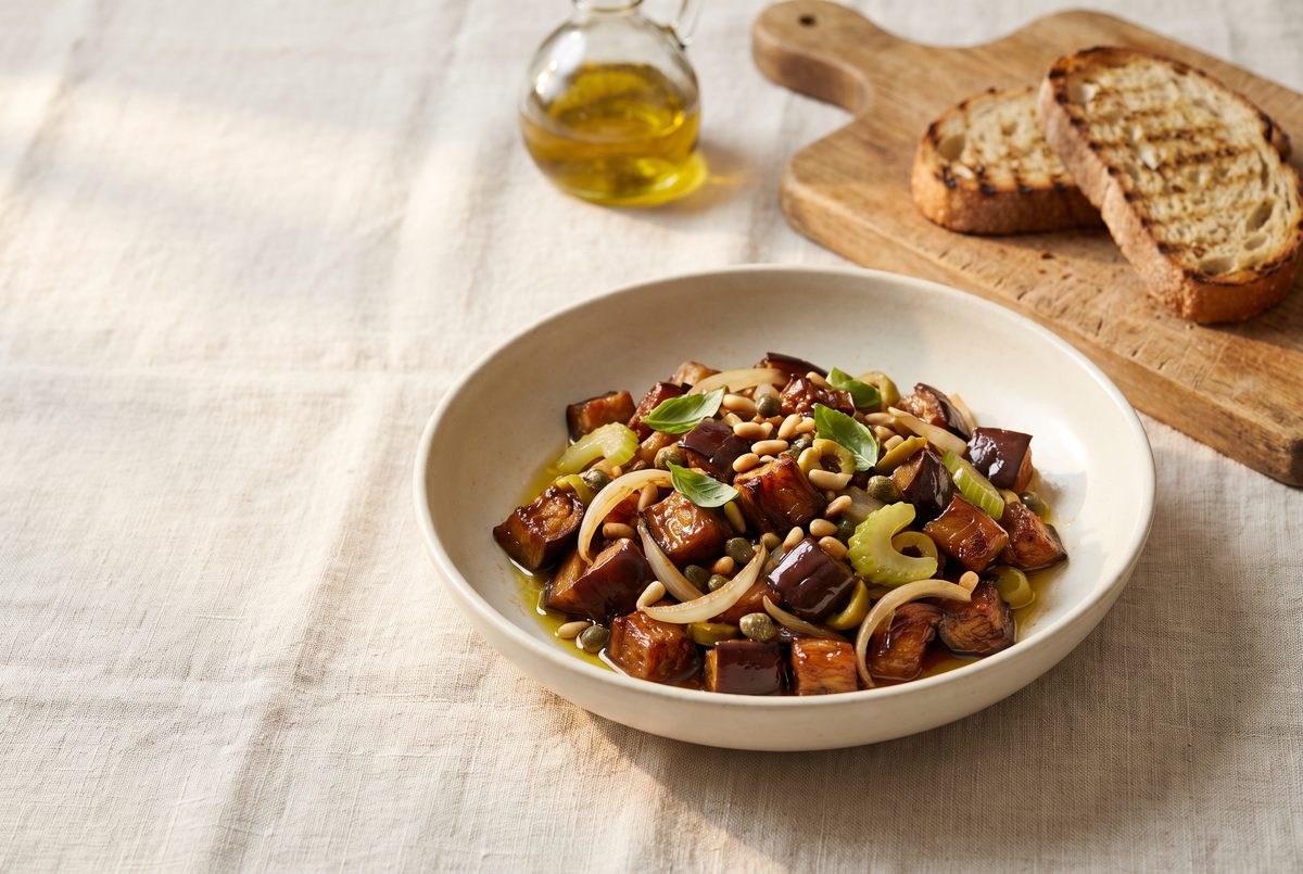 A bowl of eggplant caponata with pine nuts, capers, onions, and celery, served with grilled bread and olive oil on a light tablecloth.