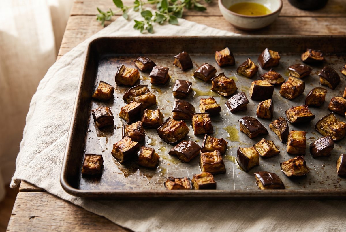 Dark, wrinkled roasted eggplant cubes spread across a plain black sheet pan