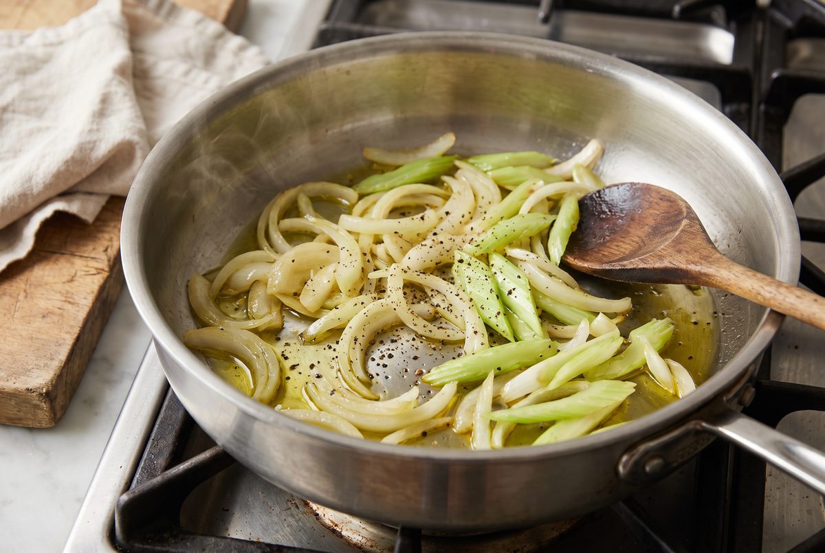 Sliced onion and diagonal-cut celery softening in olive oil in a wide stainless-steel pan