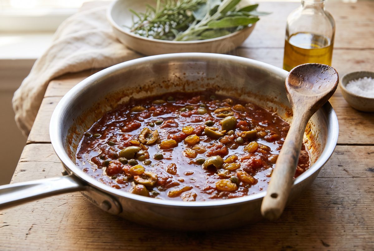 Glossy dark tomato base with olives and capers in a wide pan, wooden spoon resting against the rim