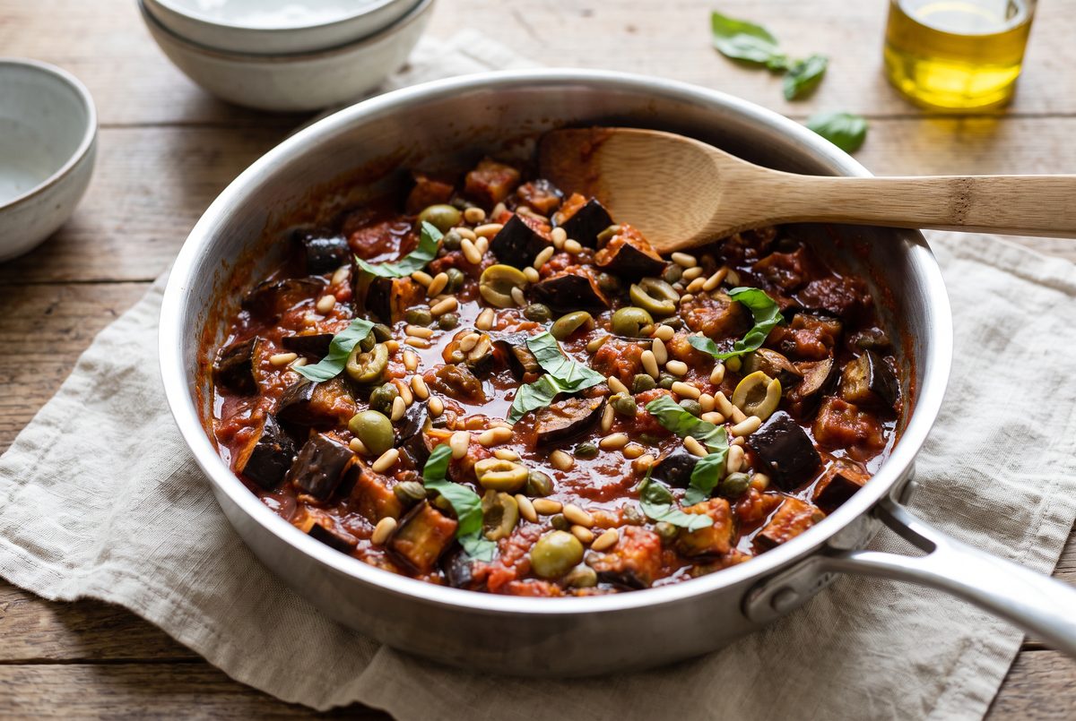 Roasted eggplant cubes just combined with the tomato, olives and pine nuts in a wide pan, flat wooden spoon resting on the rim