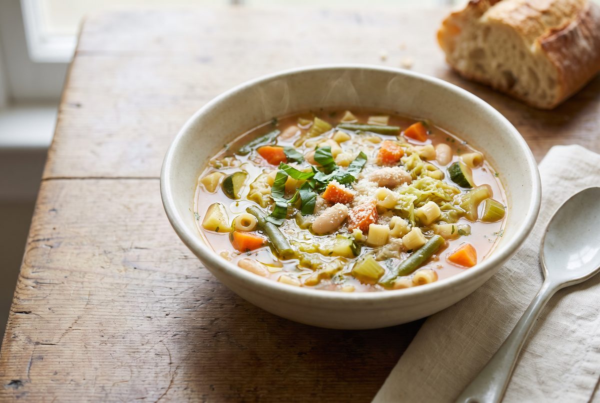 A bowl of minestrone soup with vegetables, pasta, and beans, garnished with fresh herbs and grated cheese, sits on a rustic wooden table next to a piece of crusty bread and a spoon.