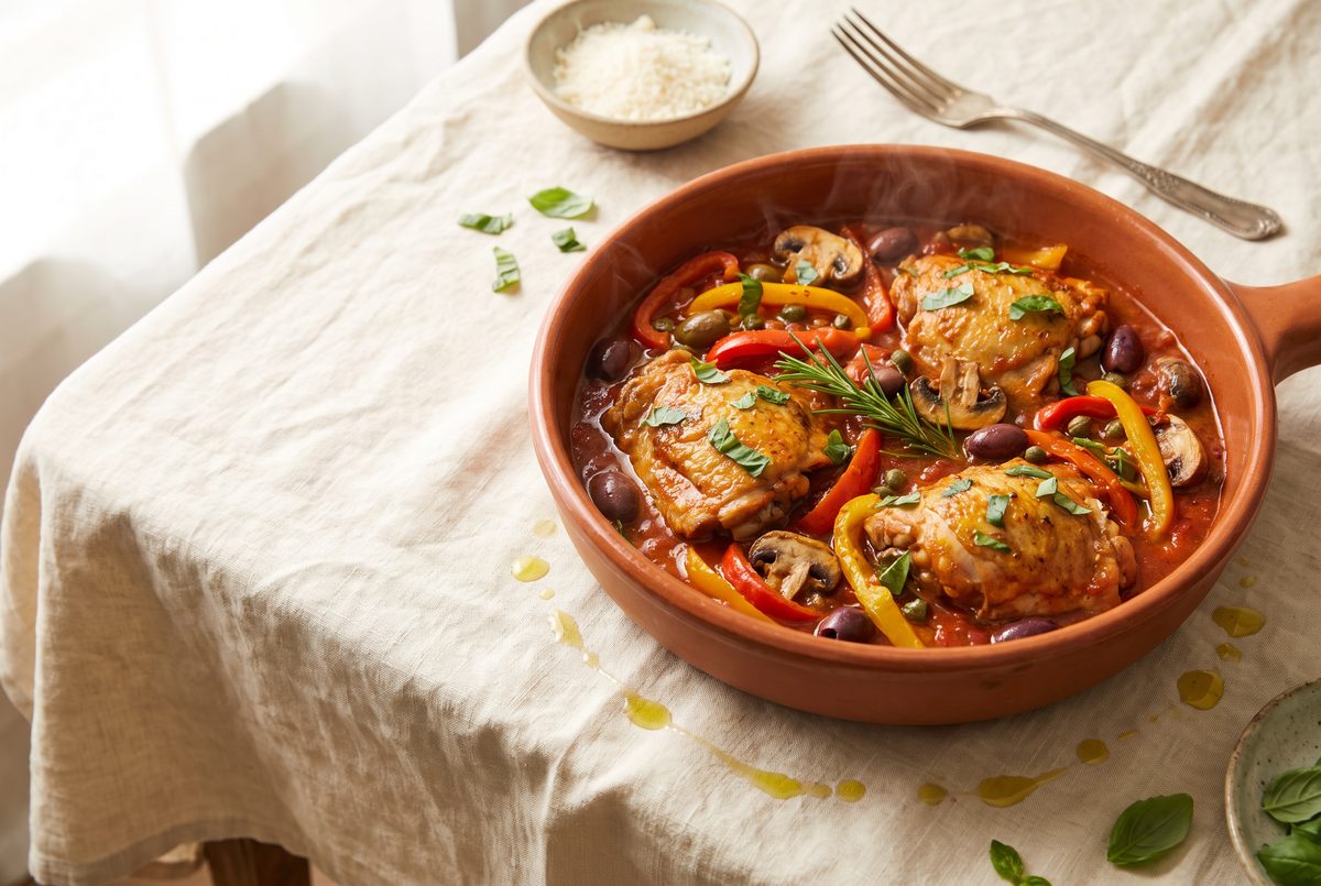 A terracotta dish of chicken cacciatore with bell peppers, mushrooms, olives, and herbs is presented on a light tablecloth.