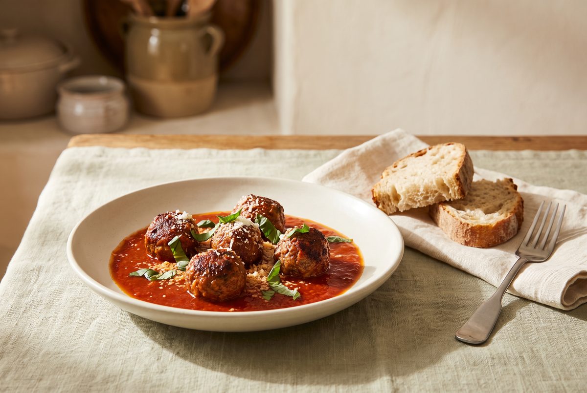 A plate of meatballs in tomato sauce, garnished with basil and grated cheese, is served with slices of bread and a fork on a linen tablecloth.