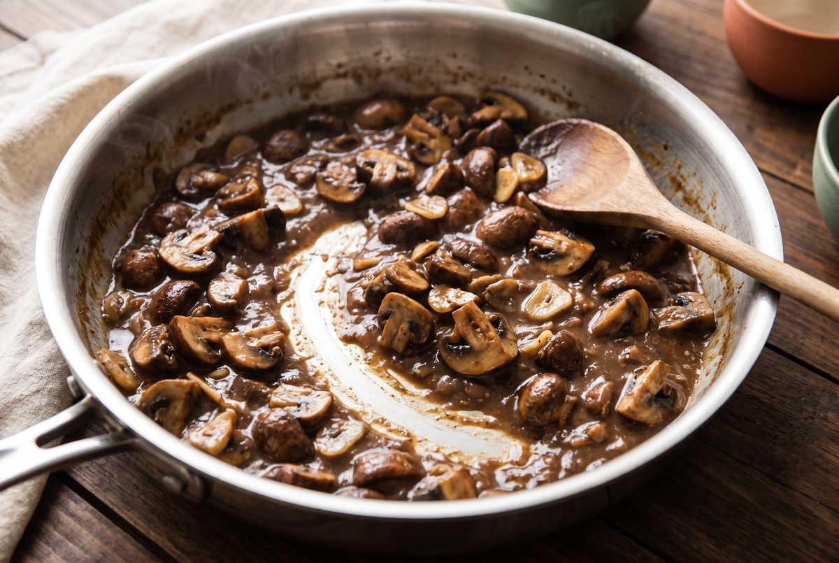 Browned mushrooms in a glossy dark brown sauce reduced in a wide stainless-steel pan, a wooden spoon resting against the side