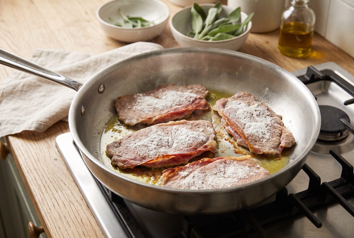 Four veal cutlets prosciutto side down in a stainless-steel pan, ham edges crisped and browned