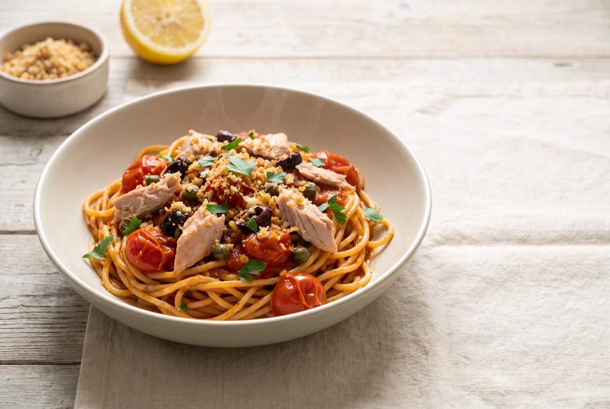 A bowl of spaghetti with tuna, roasted cherry tomatoes, black olives, capers, and breadcrumbs, garnished with parsley, on a wooden table.