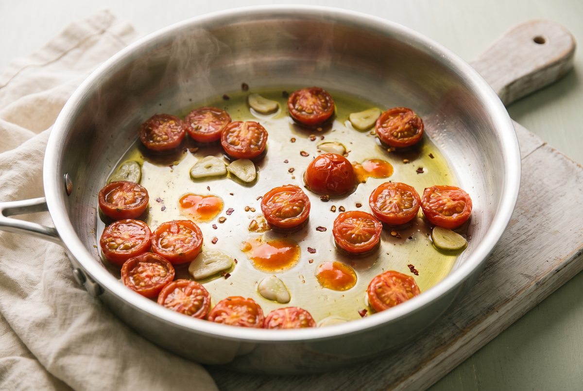 Halved cherry tomatoes collapsing in olive oil with sliced garlic and chili flakes in a wide pan