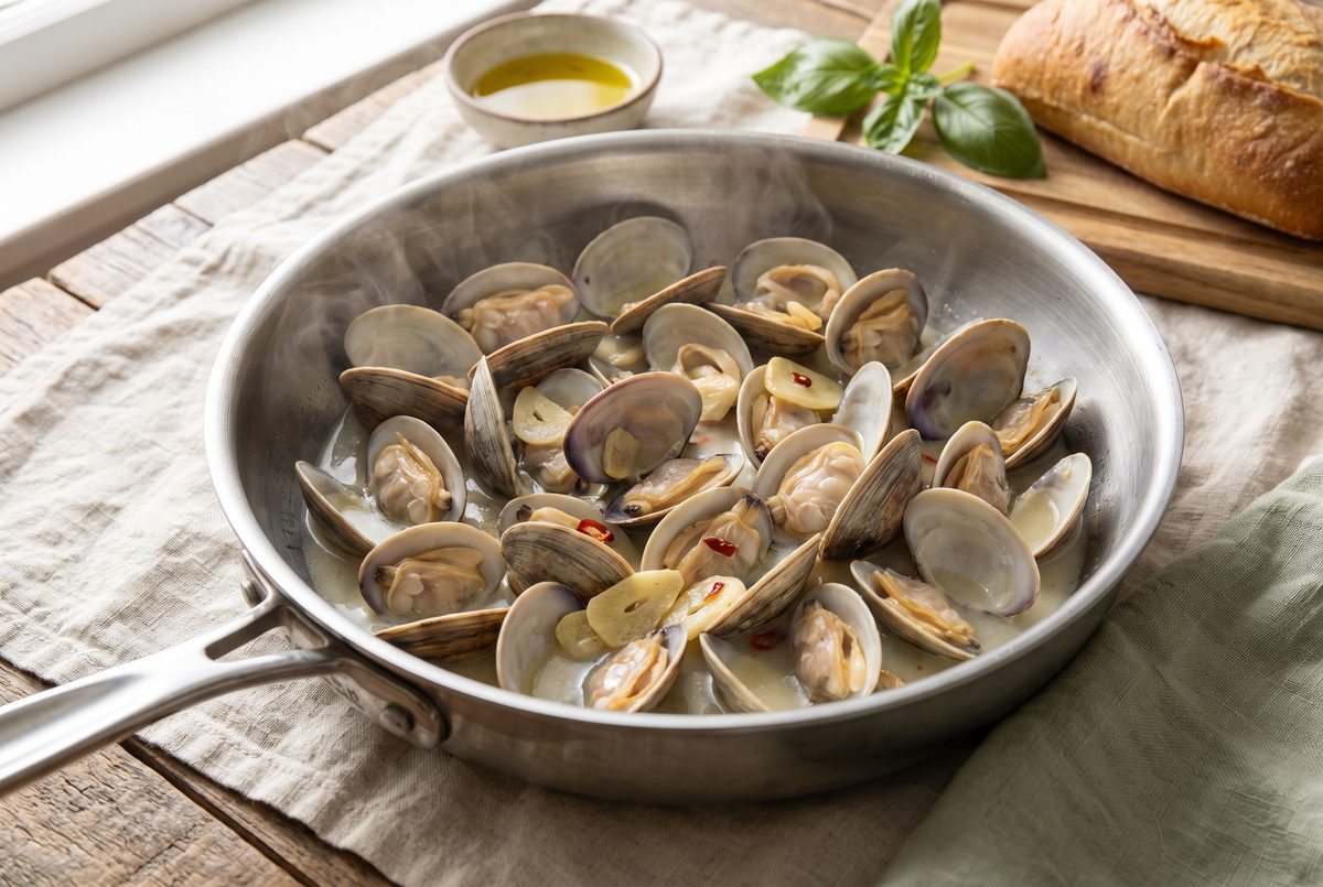 Littleneck clams wide open in a shallow stainless-steel pan with sliced garlic and a shallow pool of wine and clam liquor
